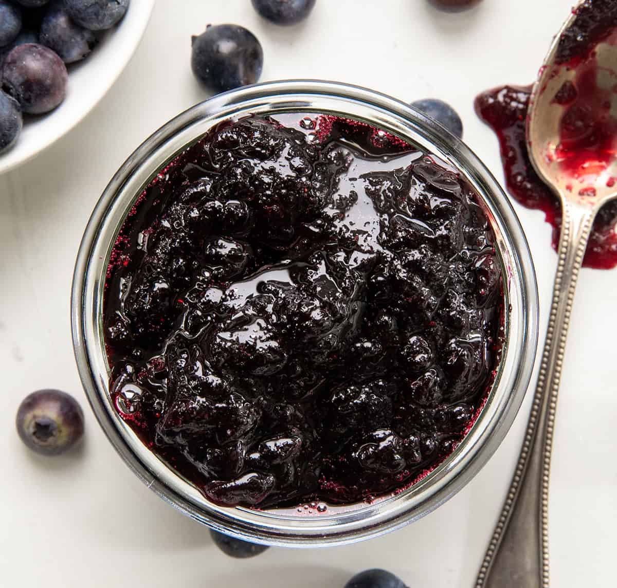 This Bourbon Blueberry Jam is thick, rich, and just sweet enough, with a warm depth of flavor from the bourbon... but does not taste like alcohol at all! Itโs made with pectin, so it sets up nicely and comes together quickly. If you have tried my blueberry jam without pectin (or bourbon) and loved it, I think youโll really enjoy this version, too! Close up of the top of a jar of Bourbon Blueberry Jam on a white table from overhead.