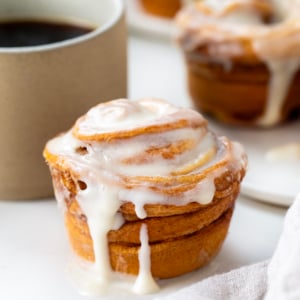 Single Cinnamon Roll Cruffin on a white table with coffee and more cruffins in the background.