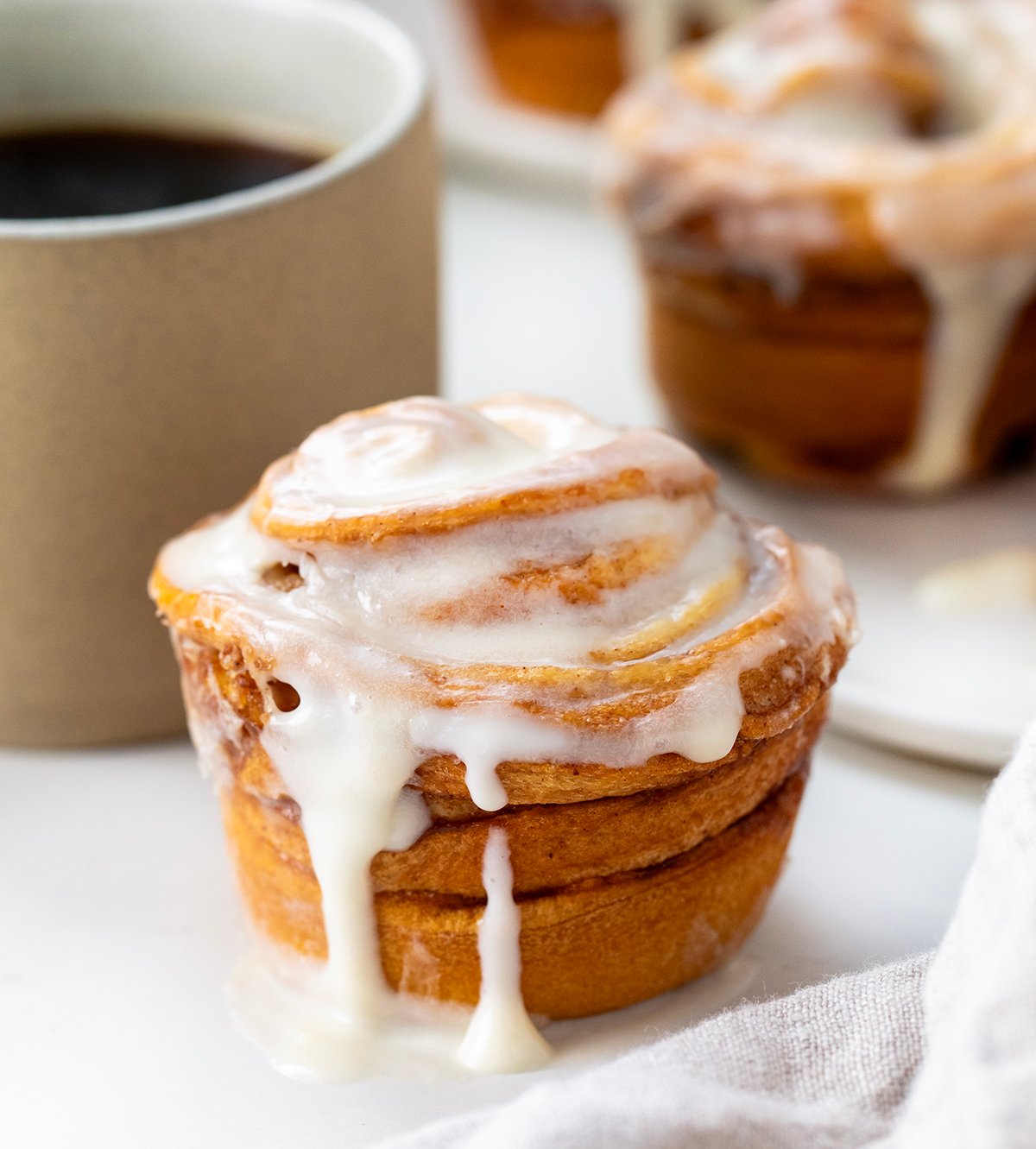 These Cinnamon Roll Cruffins are filled with a buttery cinnamon sugar mixture and baked in a muffin tin for the perfect pull-apart swirl. Each one is soft, flaky, and full of sweet cinnamon flavor! Then they are coated in a smooth cream cheese glaze that melts into every layer! Single Cinnamon Roll Cruffin on a white table with coffee and more cruffins in the background.