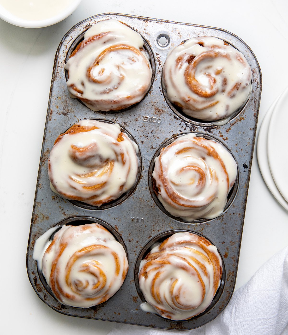 These Cinnamon Roll Cruffins are filled with a buttery cinnamon sugar mixture and baked in a muffin tin for the perfect pull-apart swirl. Each one is soft, flaky, and full of sweet cinnamon flavor! Then they are coated in a smooth cream cheese glaze that melts into every layer! Muffin pan of Cinnamon Roll Cruffins on a white table from overhead.