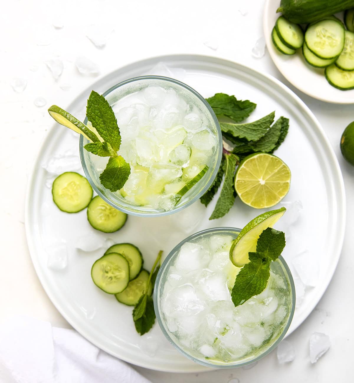 Cool down with this refreshing Cucumber Mint Refresher cocktail! Made with fresh mint, cucumber, lime juice, vodka (or gin), and bubbly club soda. Perfect for summer sipping or any time you need a light, fizzy, (almost healthy!!) drink. Glasses of Cucumber Mint Refresher on a white platter on a white table from overhead.