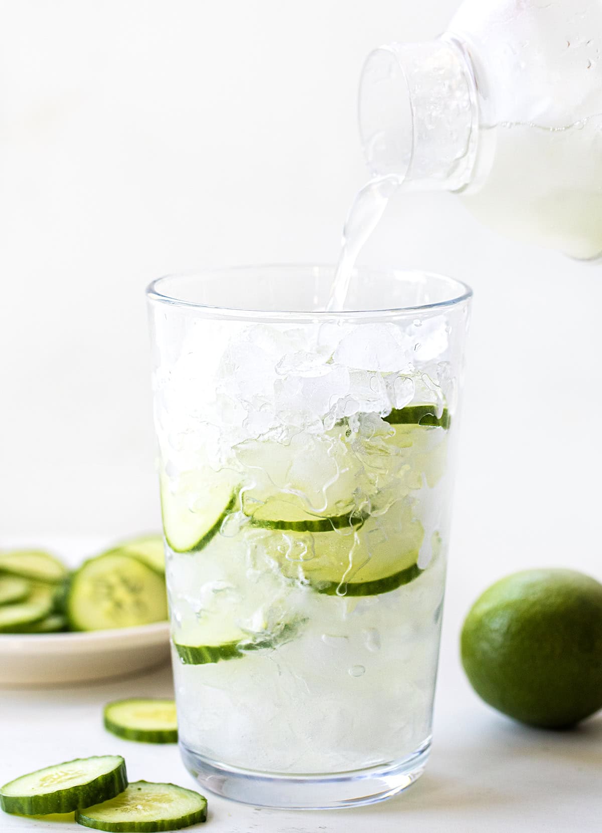 Cool down with this refreshing Cucumber Mint Refresher cocktail! Made with fresh mint, cucumber, lime juice, vodka (or gin), and bubbly club soda. Perfect for summer sipping or any time you need a light, fizzy, (almost healthy!!) drink. Pouring Cucumber Mint Refresher into a glass filled with ice on a white table.