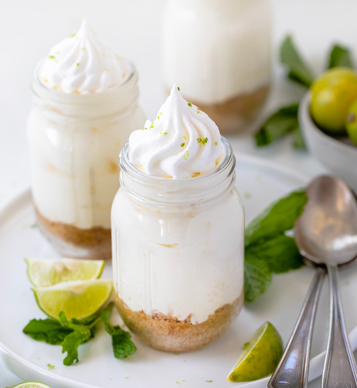 Creamy Key Lime Cheesecake Cups have all the tangy flavor of Key Lime Pie in easy, individual servings, perfect for parties and make-ahead desserts! Jars of Key Lime Cheesecake on a white platter on a white table with fresh key limes and mint.