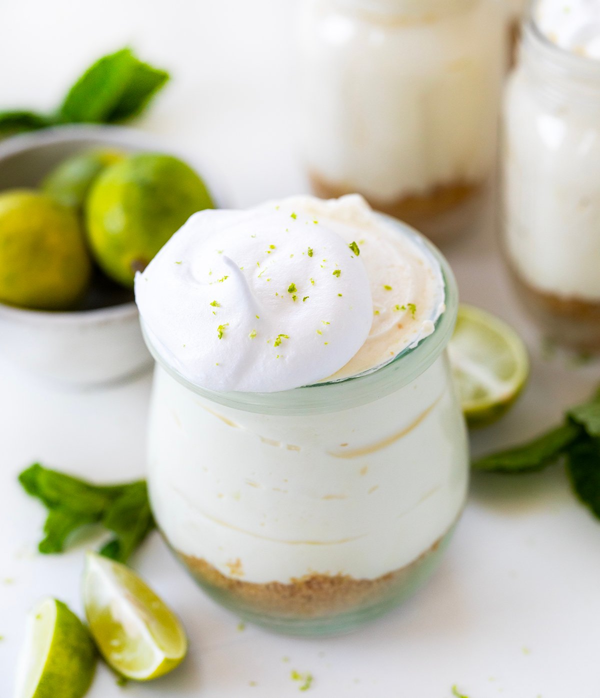 Creamy Key Lime Cheesecake Cups have all the tangy flavor of Key Lime Pie in easy, individual servings, perfect for parties and make-ahead desserts! Jars of Key Lime Cheesecake on a white platter on a white table with fresh key limes and mint.