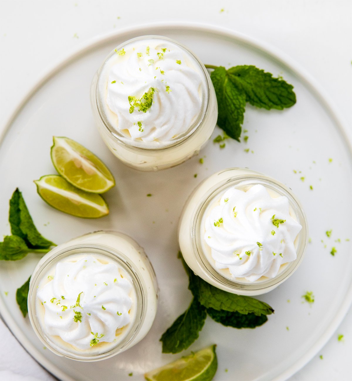 Creamy Key Lime Cheesecake Cups have all the tangy flavor of Key Lime Pie in easy, individual servings, perfect for parties and make-ahead desserts! Jars of Key Lime Cheesecake on a white platter on a white table with fresh key limes and mint from overhead.