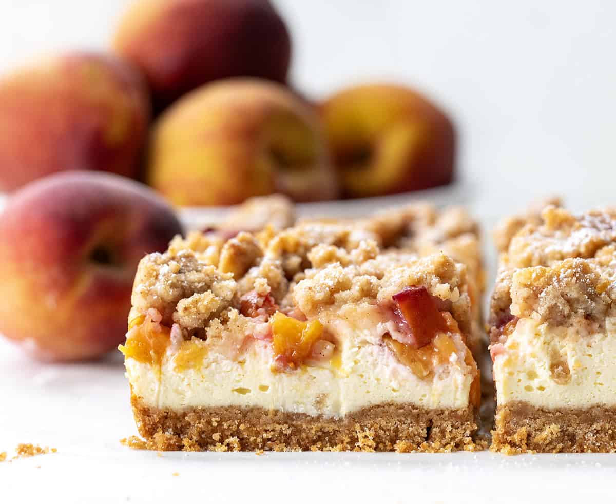 These Peach Cheesecake Bars have a buttery graham cracker crust, a creamy cheesecake layer, sweet diced peaches, and a cinnamon crumble topping. Every bite tastes like peach cobbler and cheesecake combined! Close up of Peach Cobbler Cheesecake Bars on a white table with peaches in the bakcground.