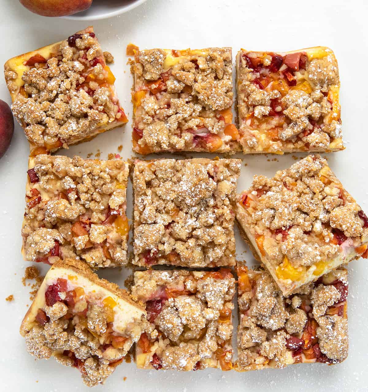 These Peach Cheesecake Bars have a buttery graham cracker crust, a creamy cheesecake layer, sweet diced peaches, and a cinnamon crumble topping. Every bite tastes like peach cobbler and cheesecake combined! Peach Cobbler Cheesecake Bars cut into squares and a couple of them tilted on their side on a white table from overhead.