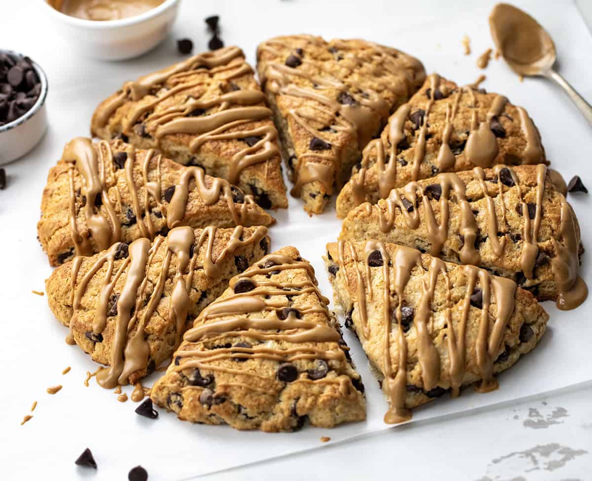 Peanut Butter Chocolate Chip Scones are my buttery, flaky chocolate chip scones taken to the next level with peanut butter mixed into the dough and drizzled on top. They are perfect for all my chocolate peanut butter lovers out there! Peanut Butter Chocolate Chip Scones in a circle on a white table drizzled with peanut butter.