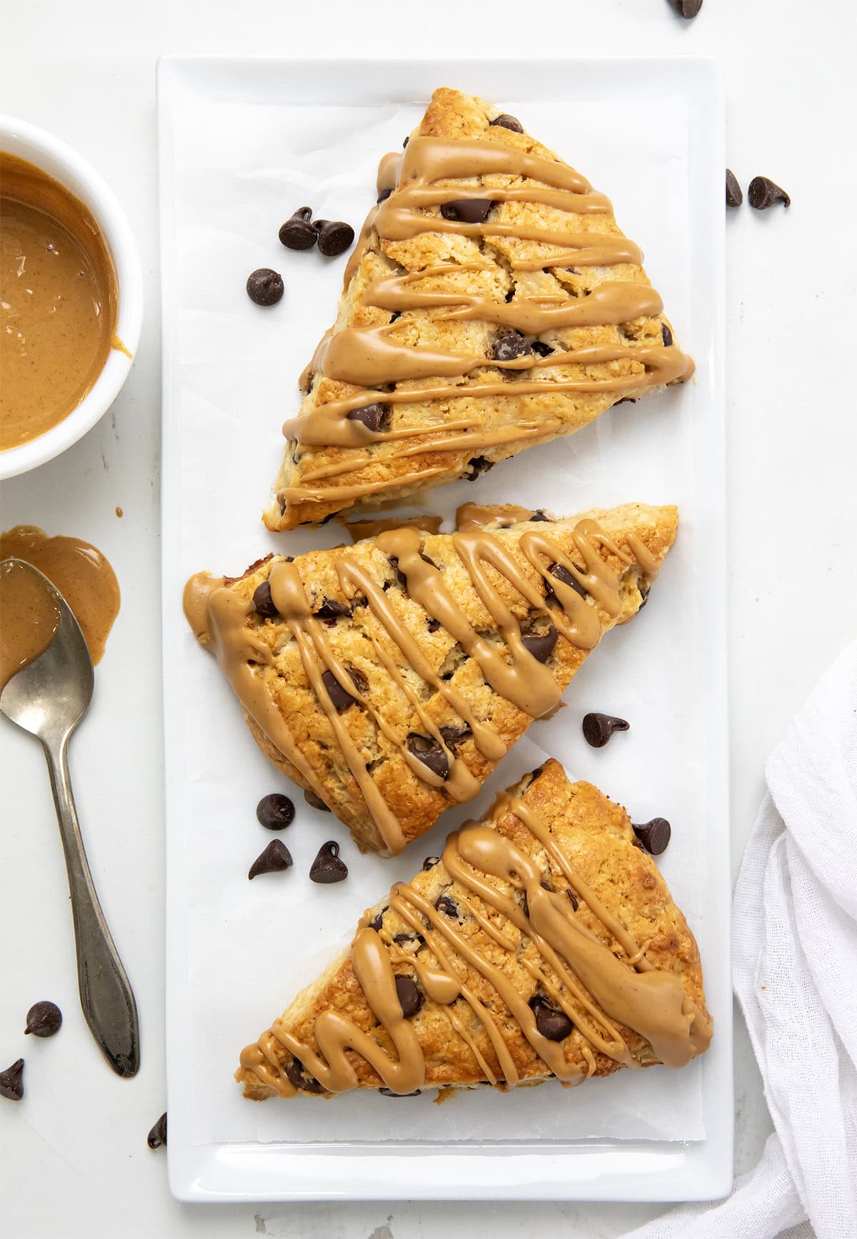 Peanut Butter Chocolate Chip Scones are my buttery, flaky chocolate chip scones taken to the next level with peanut butter mixed into the dough and drizzled on top. They are perfect for all my chocolate peanut butter lovers out there! Peanut Butter Chocolate Chip Scones on a white platter from overhead.