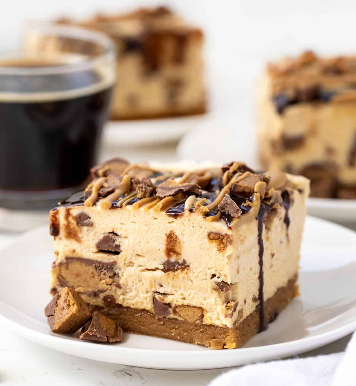 These Peanut Butter Cup Cheesecake Bars take my peanut butter cheesecake (which I absolutely love!) and turn it into easy-to-serve dessert bars. The bars start with a homemade easy peanut butter cookie crust, topped with a creamy peanut butter cheesecake filling loaded with chopped peanut butter cups. Peanut Butter Cup Cheesecake Bars on a white plate on a white table.