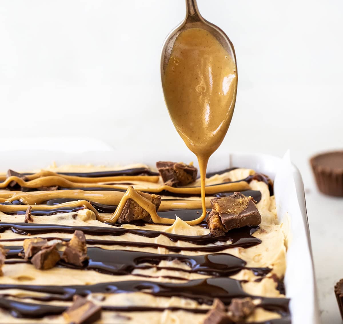 These Peanut Butter Cup Cheesecake Bars take my peanut butter cheesecake (which I absolutely love!) and turn it into easy-to-serve dessert bars. The bars start with a homemade easy peanut butter cookie crust, topped with a creamy peanut butter cheesecake filling loaded with chopped peanut butter cups. Drizzling peanut butter over Peanut Butter Cup Cheesecake Bars.