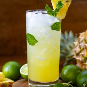One Pineapple Mojito on a wooden table surrounded by limes and pineapple.
