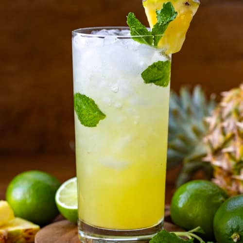 One Pineapple Mojito on a wooden table surrounded by limes and pineapple.