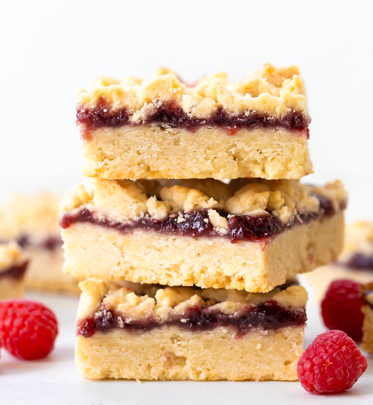 Raspberry Almond Bars are a sweet and simple treat with a buttery almond shortbread dough doing double duty as both the crust and the crumbly topping, with a layer of raspberry jam in between. Just because they are so EASY to make doesn't mean they lack flavor! Stack of Raspberry Almond Bars on a white table.