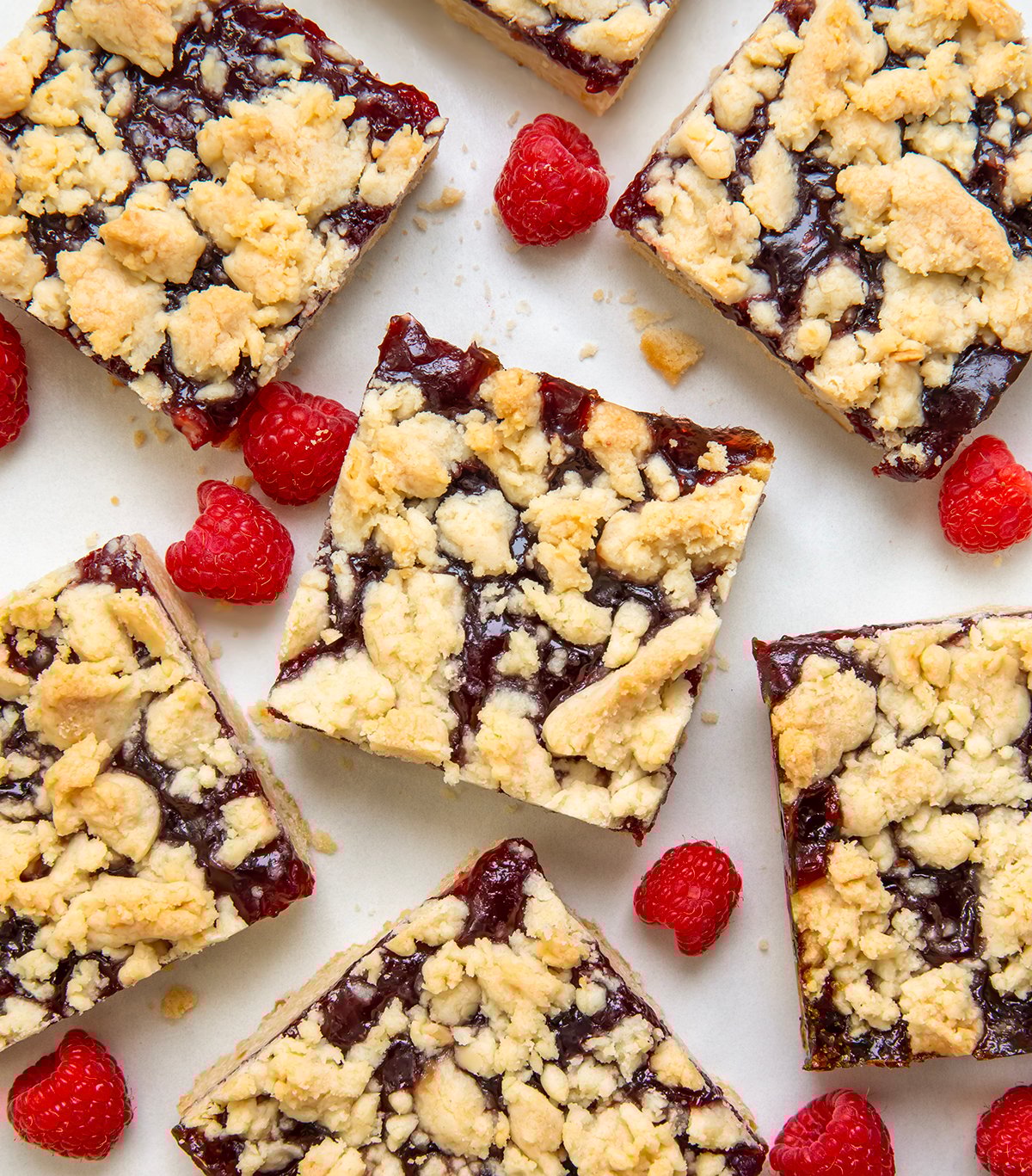 Raspberry Almond Bars are a sweet and simple treat with a buttery almond shortbread dough doing double duty as both the crust and the crumbly topping, with a layer of raspberry jam in between. Just because they are so EASY to make doesn't mean they lack flavor! Raspberry Almond Bars cut into squares on a white tale from overhead.
