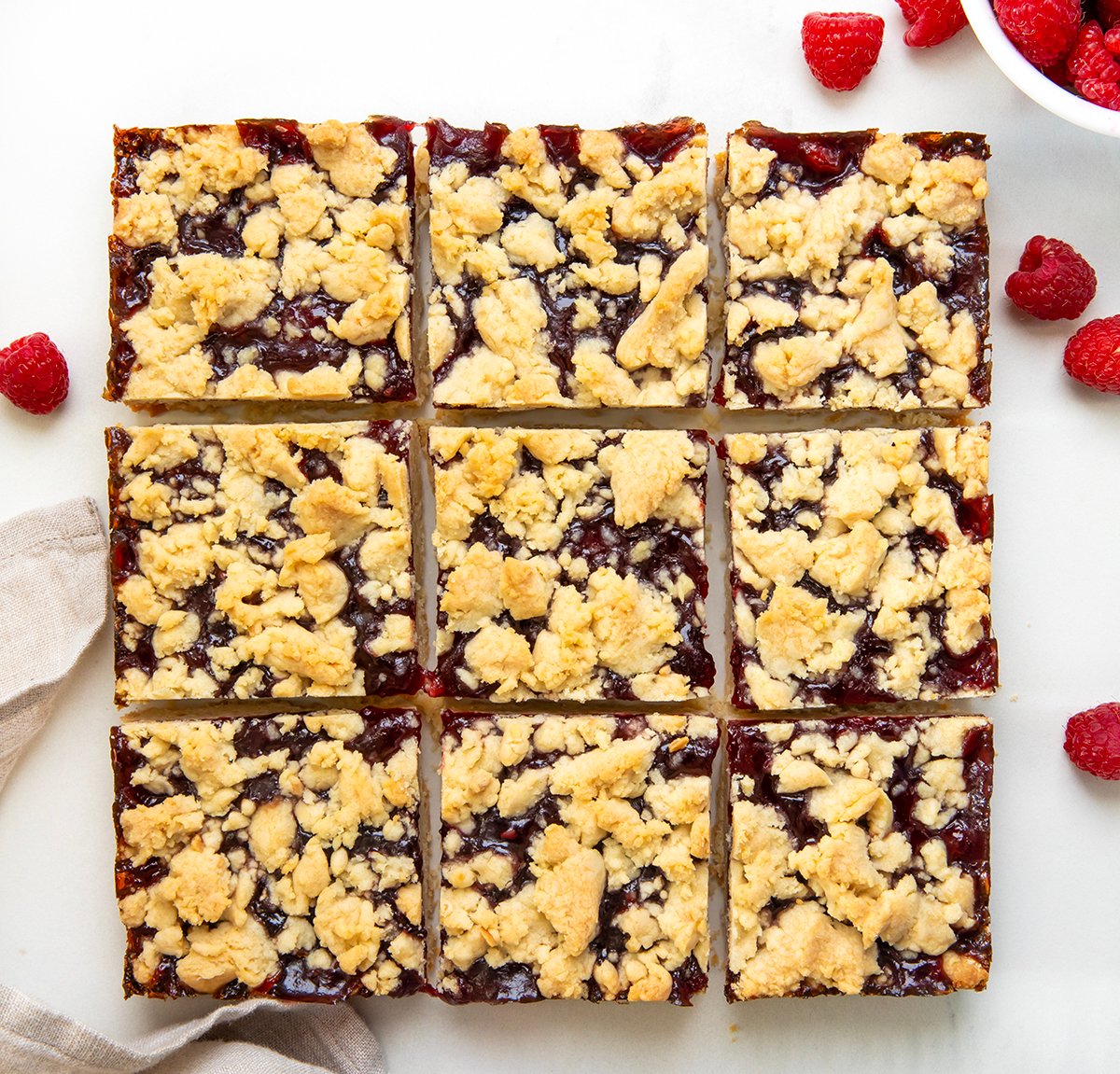 Raspberry Almond Bars are a sweet and simple treat with a buttery almond shortbread dough doing double duty as both the crust and the crumbly topping, with a layer of raspberry jam in between. Just because they are so EASY to make doesn't mean they lack flavor! Raspberry Almond Bars cut into squares on a white tale from overhead.