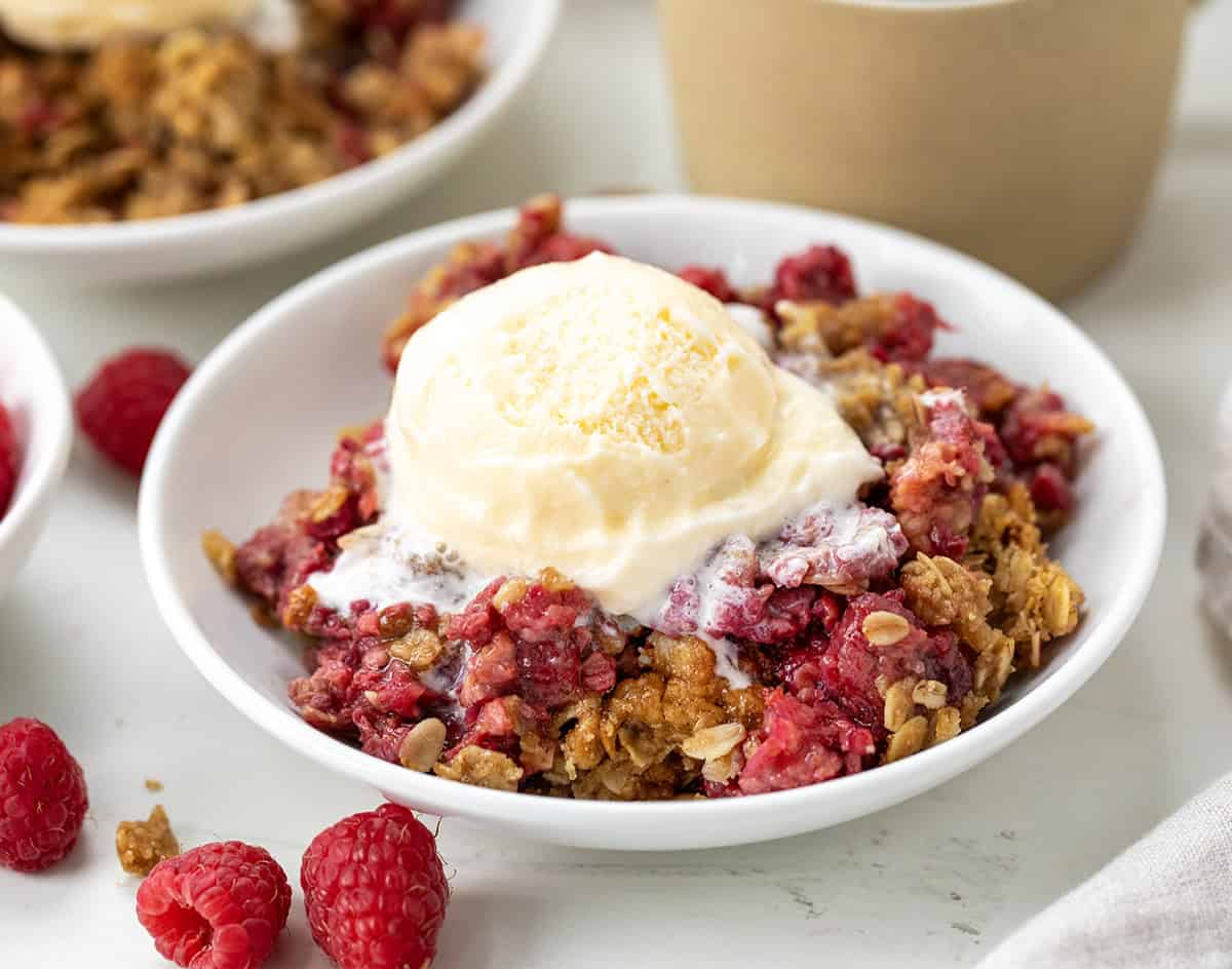 If youโre looking for an easy raspberry dessert, you have got to try this Raspberry Crisp! Itโs made with fresh raspberries and topped with a buttery oat crumble that bakes up golden and crisp. So simple, so good, and even better with a scoop of vanilla ice cream on top! Plate of Raspberry Crisp with a scoop of ice cream on a white table.