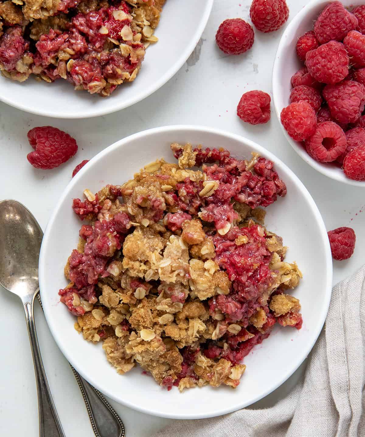 If youโre looking for an easy raspberry dessert, you have got to try this Raspberry Crisp! Itโs made with fresh raspberries and topped with a buttery oat crumble that bakes up golden and crisp. So simple, so good, and even better with a scoop of vanilla ice cream on top! Plates of Raspberry Crisp on a white table with fresh raspberries from overhead.
