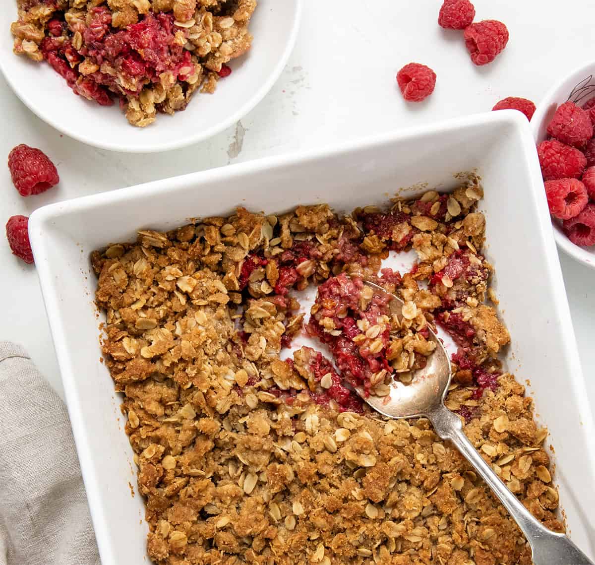 If youโre looking for an easy raspberry dessert, you have got to try this Raspberry Crisp! Itโs made with fresh raspberries and topped with a buttery oat crumble that bakes up golden and crisp. So simple, so good, and even better with a scoop of vanilla ice cream on top! Pan of Raspberry Crisp with a spoon in it on a white table from overhead.