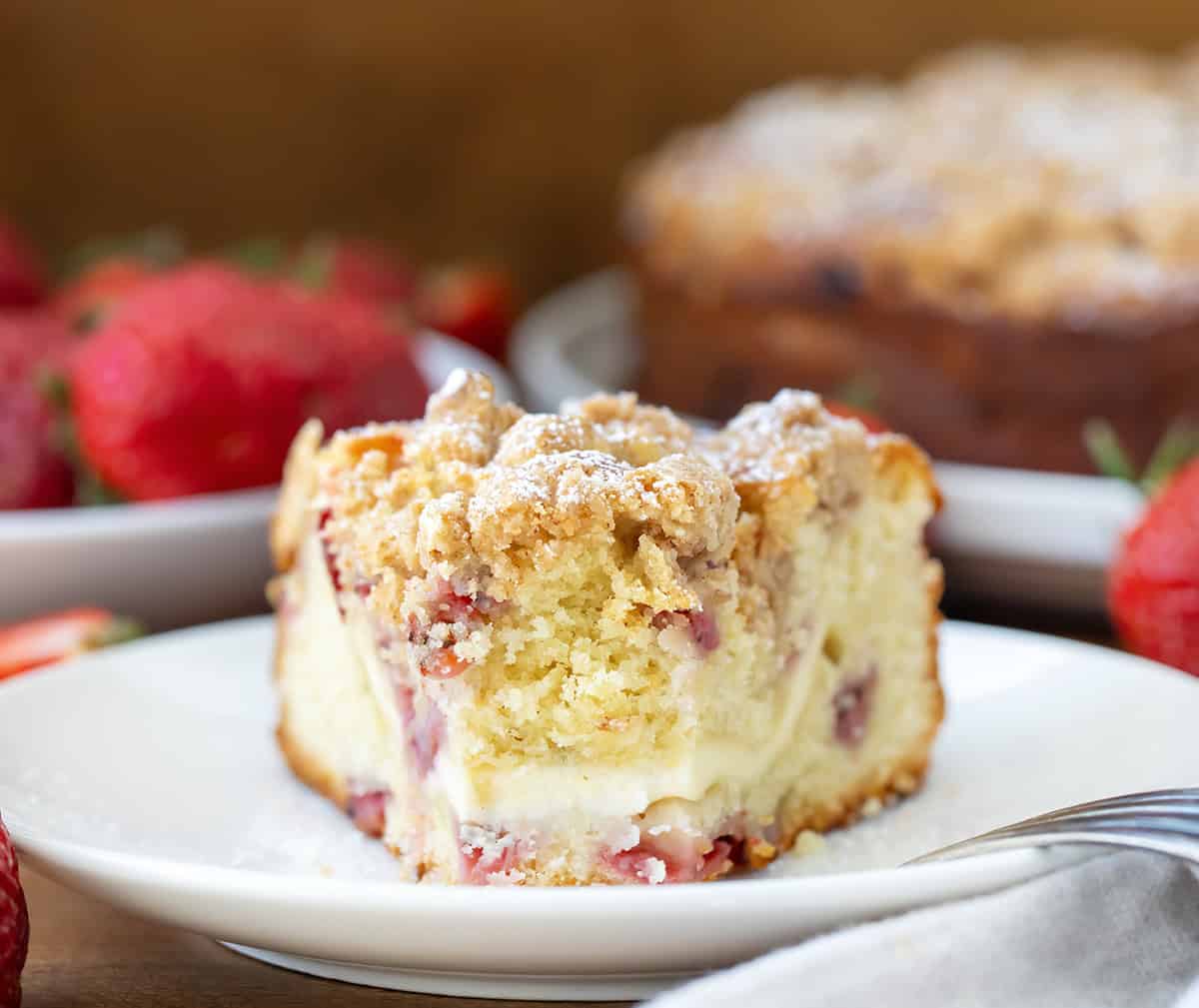 This Strawberries and Cream Coffee Cake is soft, sweet, and full of fresh strawberry flavor. It has layers of fluffy vanilla cake, a creamy cheesecake filling, and a buttery crumble on top. Every bite is packed with juicy strawberries! You can enjoy a slice of this cake for breakfast, brunch, dessert, or any time of the day! Slice of Strawberries and Cream Coffee Cake on a plate with a bite removed and the fork resting on the plate.