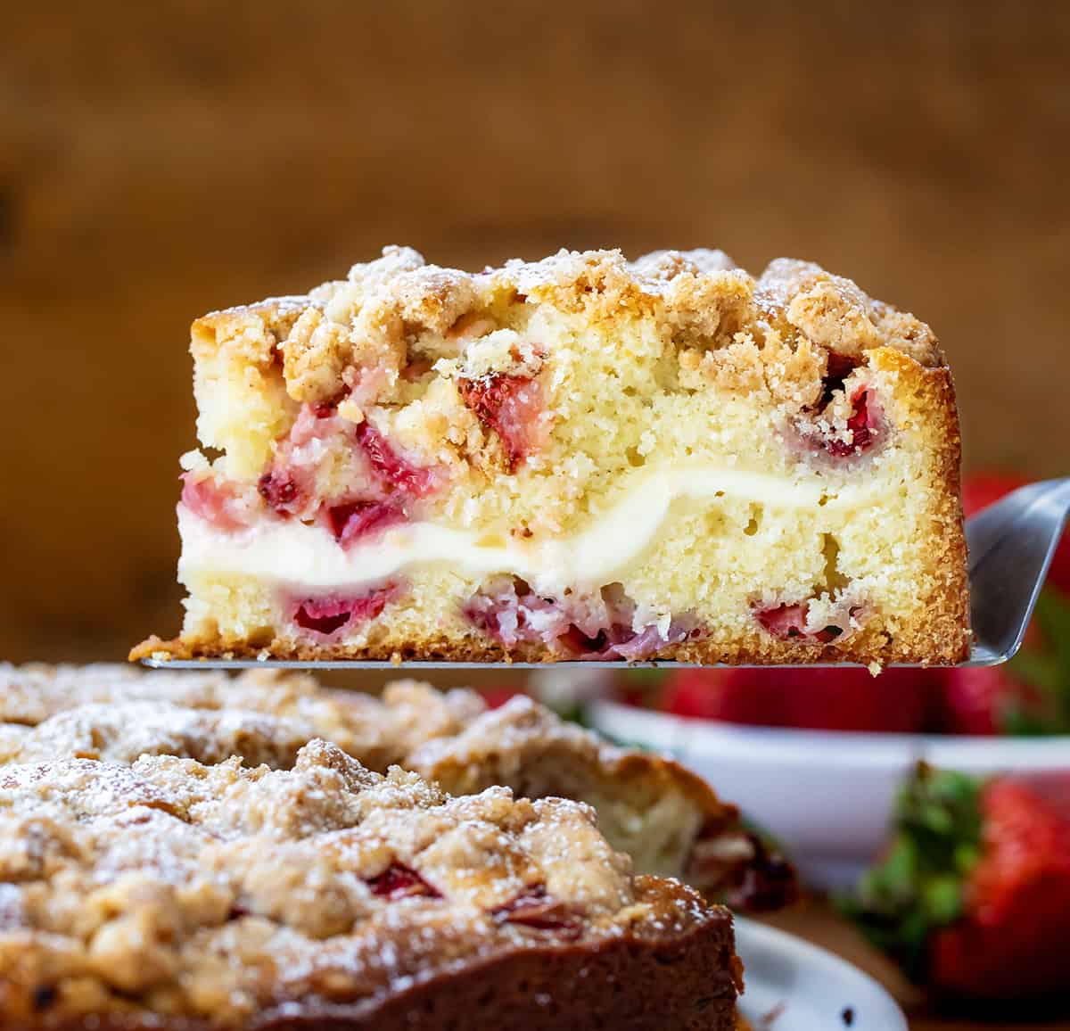 This Strawberries and Cream Coffee Cake is soft, sweet, and full of fresh strawberry flavor. It has layers of fluffy vanilla cake, a creamy cheesecake filling, and a buttery crumble on top. Every bite is packed with juicy strawberries! You can enjoy a slice of this cake for breakfast, brunch, dessert, or any time of the day! Picking up a slice of Strawberries and Cream Coffee Cake from the serving platter.