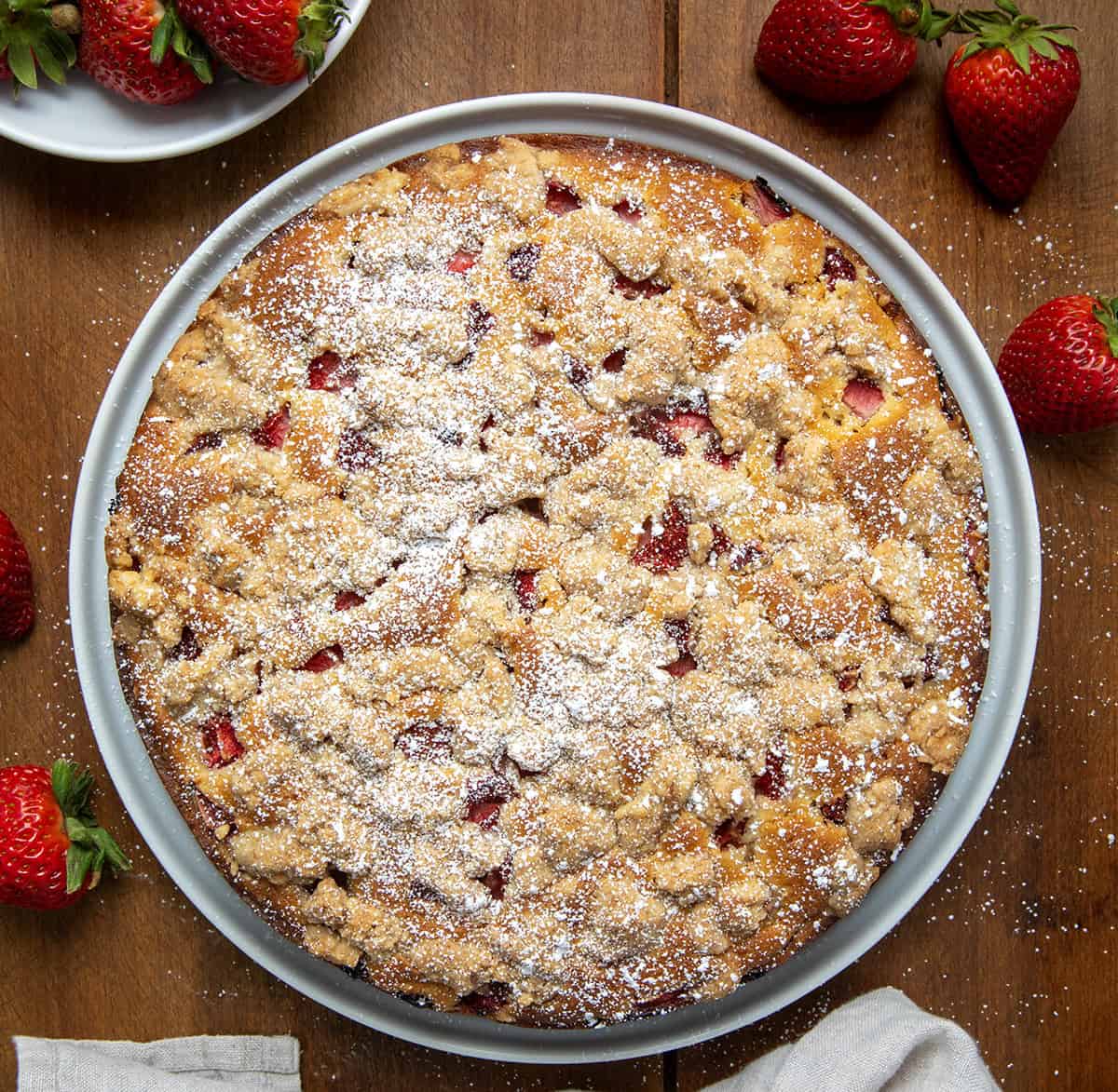 This Strawberries and Cream Coffee Cake is soft, sweet, and full of fresh strawberry flavor. It has layers of fluffy vanilla cake, a creamy cheesecake filling, and a buttery crumble on top. Every bite is packed with juicy strawberries! You can enjoy a slice of this cake for breakfast, brunch, dessert, or any time of the day! Whole Strawberries and Cream Coffee Cake on a white serving plate on a wooden table from overhead.
