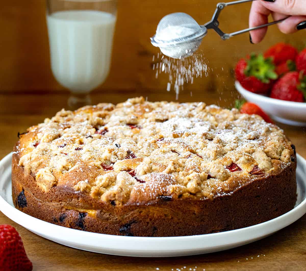 This Strawberries and Cream Coffee Cake is soft, sweet, and full of fresh strawberry flavor. It has layers of fluffy vanilla cake, a creamy cheesecake filling, and a buttery crumble on top. Every bite is packed with juicy strawberries! You can enjoy a slice of this cake for breakfast, brunch, dessert, or any time of the day! Dusting Strawberries and Cream Coffee Cake with confectioners sugar.