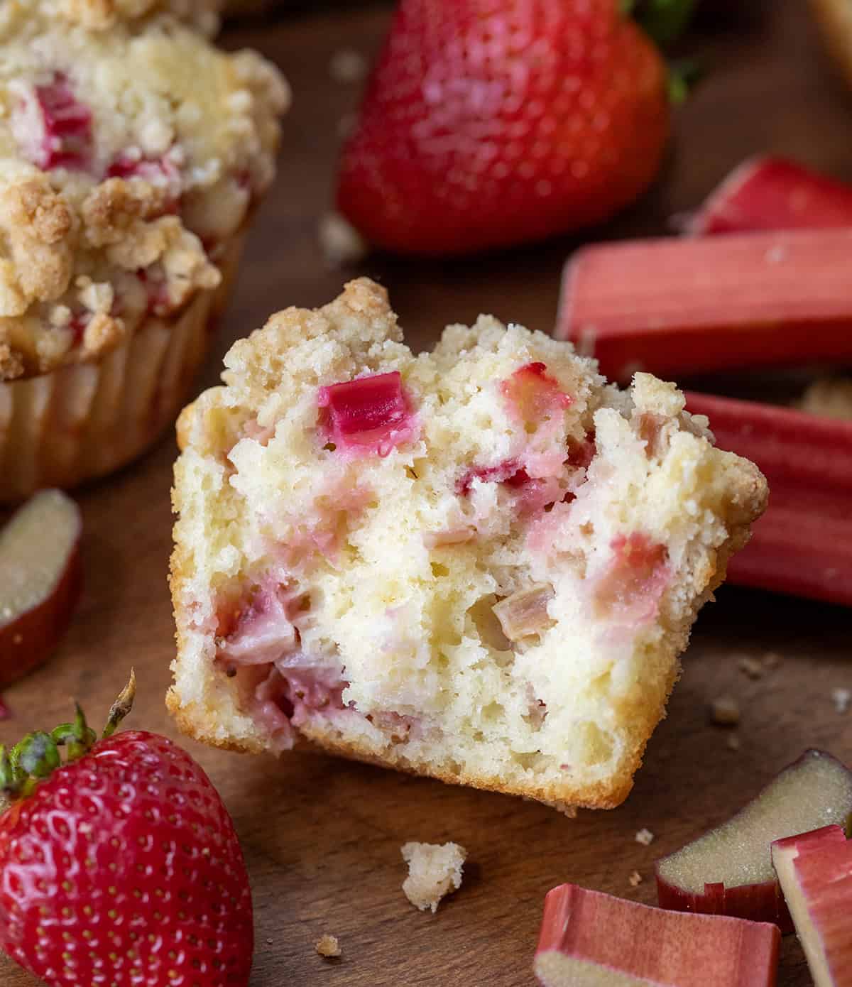 Strawberry Rhubarb Muffins are the perfect sweet-tart treat for spring and summer! Each soft, buttery muffin is packed with fresh strawberries and rhubarb, then topped with a golden crumb! Half of a Strawberry Rhubarb Muffin on a wooden table showing tender inside texture.