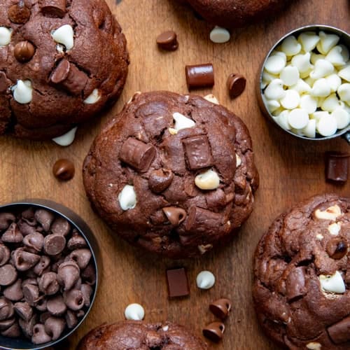 Triple Chocolate Chunk Cookies laying flat on a wooden table with chocolate chunks and chips surrounding it.