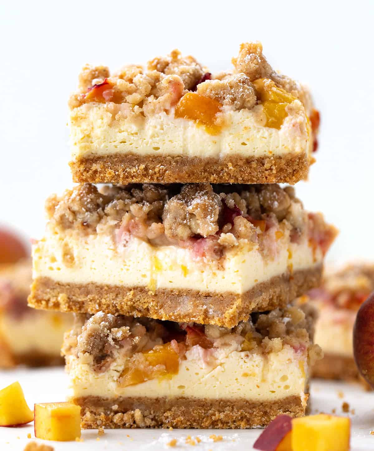 These Peach Cheesecake Bars have a buttery graham cracker crust, a creamy cheesecake layer, sweet diced peaches, and a cinnamon crumble topping. Every bite tastes like peach cobbler and cheesecake combined! Stack of peach cobbler cheesecake bars on a white table.