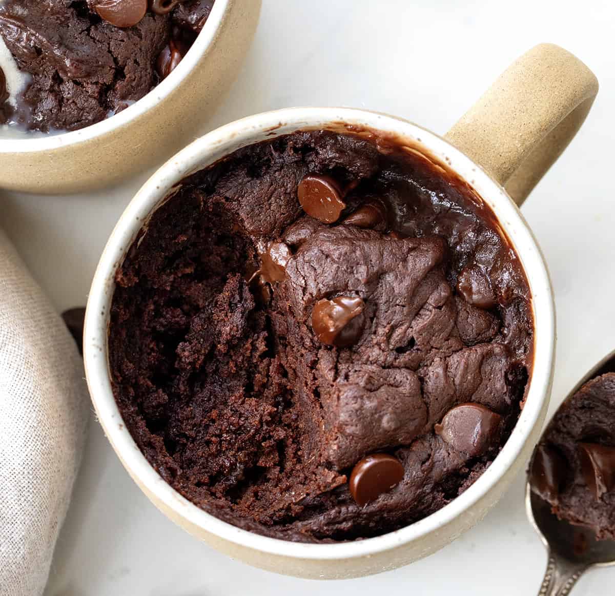 If you are craving a warm, fudgy brownie but don't want to make a whole pan, this microwave Brownie in a Mug is the answer! Itโs rich and chocolatey and comes together in just a few minutes with simple pantry staples. I make it whenever I need a quick single-serve dessert and something sweet in a hurry. No oven, no fuss, and barely any cleanup! Brownie In A Mug on a white table from overhead showing a portion has been removed and the tender texture inside.