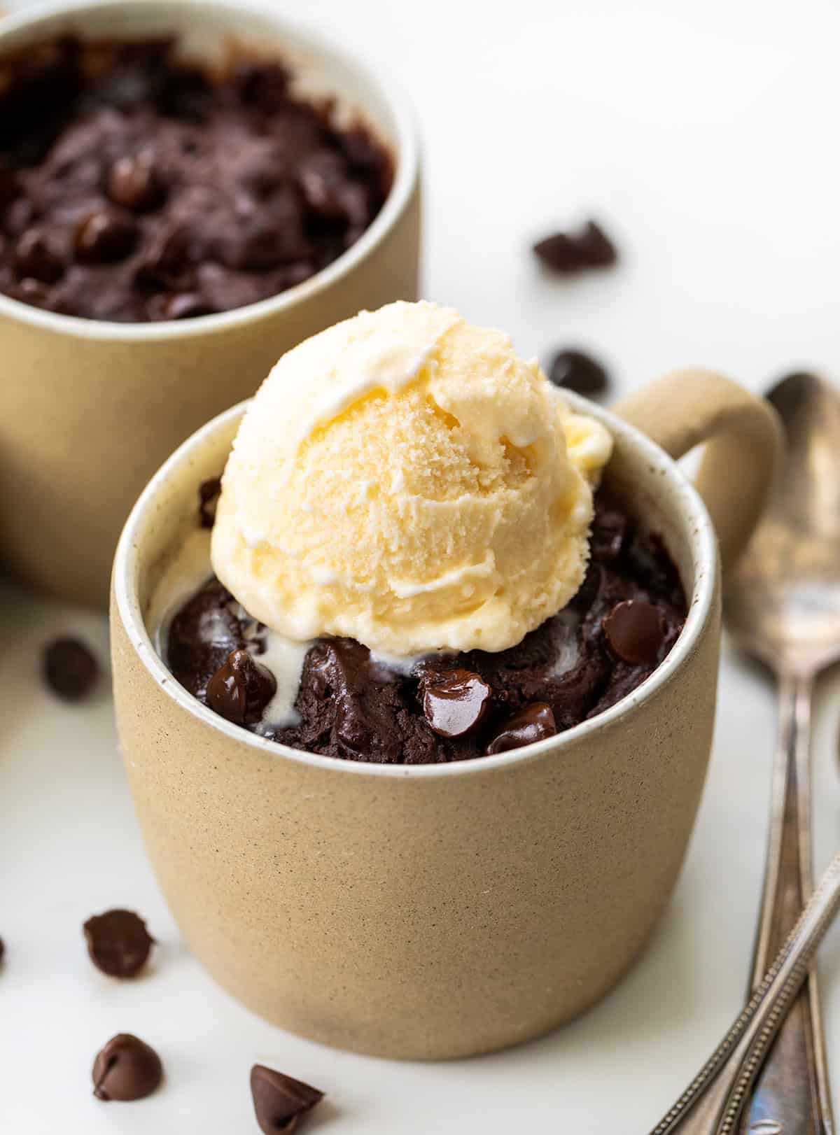 If you are craving a warm, fudgy brownie but don't want to make a whole pan, this microwave Brownie in a Mug is the answer! Itโs rich and chocolatey and comes together in just a few minutes with simple pantry staples. I make it whenever I need a quick single-serve dessert and something sweet in a hurry. No oven, no fuss, and barely any cleanup! Brownies In A Mug on a white table with spoons and one mug has ice cream scooped on top.