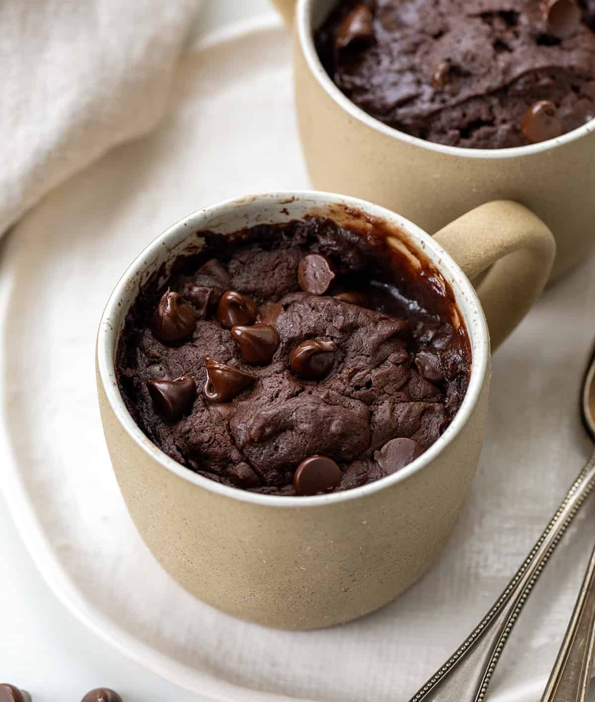 If you are craving a warm, fudgy brownie but don't want to make a whole pan, this microwave Brownie in a Mug is the answer! Itโs rich and chocolatey and comes together in just a few minutes with simple pantry staples. I make it whenever I need a quick single-serve dessert and something sweet in a hurry. No oven, no fuss, and barely any cleanup! Brownie In A Mug on a white platter on a white table with spoons.