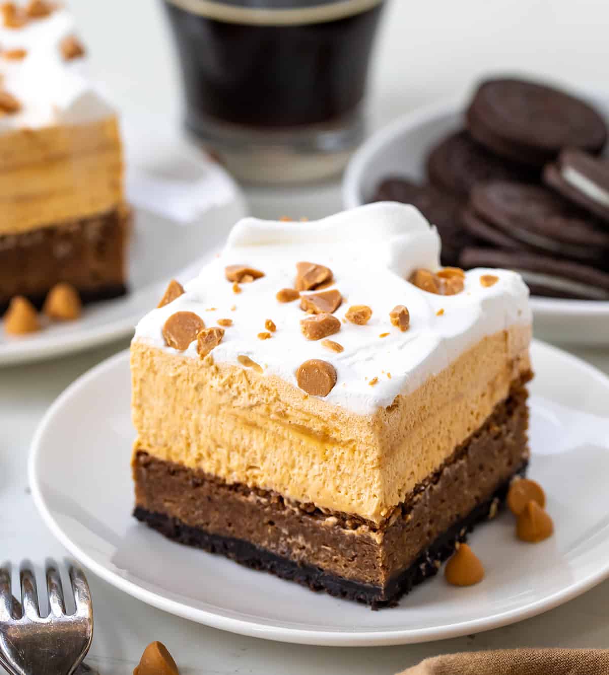 These Butterscotch Mud Bars have an Oreo cookie crust with a fudgy homemade brownie layer, topped with a silky smooth butterscotch mousse topping. I could eat each one of these layers as a dessert on its own, but putting them together creates a triple threat (in the best way!) of a dessert! One Butterscotch Mud Bar on a white plate with another bar in the background and oreos and coffee.