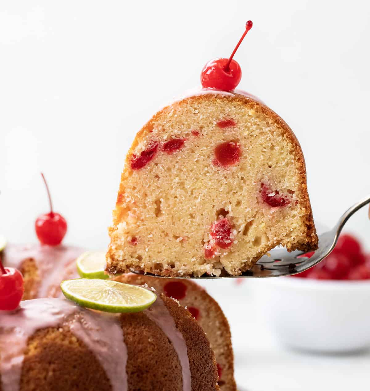 This Cherry Limeade Pound Cake is bursting with bright lime zest and sweet maraschino cherries, just like my favorite cherry limeade drink, but in cake form! I love how moist and tender it turns out! Picking up one piece of Cherry Limeade Pound Cake from the cake plate.