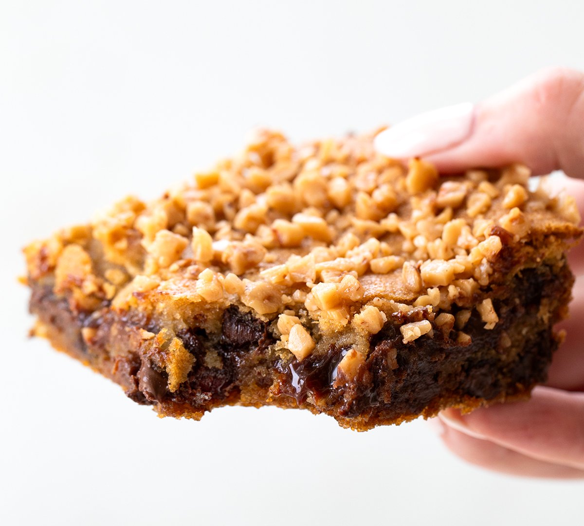 These Chocolate Chip Toffee Fudge Bars are rich, gooey, and packed with flavor! Made from my popular Hot Fudge Chocolate Chip Cookie Bars, this version adds sweet and crunchy English toffee bits for even more texture and buttery flavor! Hand holding a Chocolate Chip Toffee Fudge Bar.