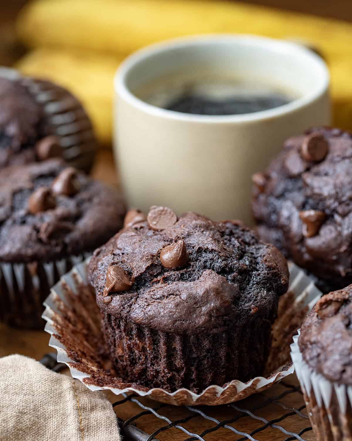 If you are looking for an easy, delicious treat that combines the rich goodness of chocolate with the natural sweetness of ripe bananas, these Double Chocolate Banana Muffins are exactly what you need. (Yes, need!) Double Chocolate Banana Muffins on a wooden table with a cup of coffee and bananas.