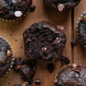 Double Chocolate Banana Muffins on a wooden table and the center muffin is cut in half showing inside texture.