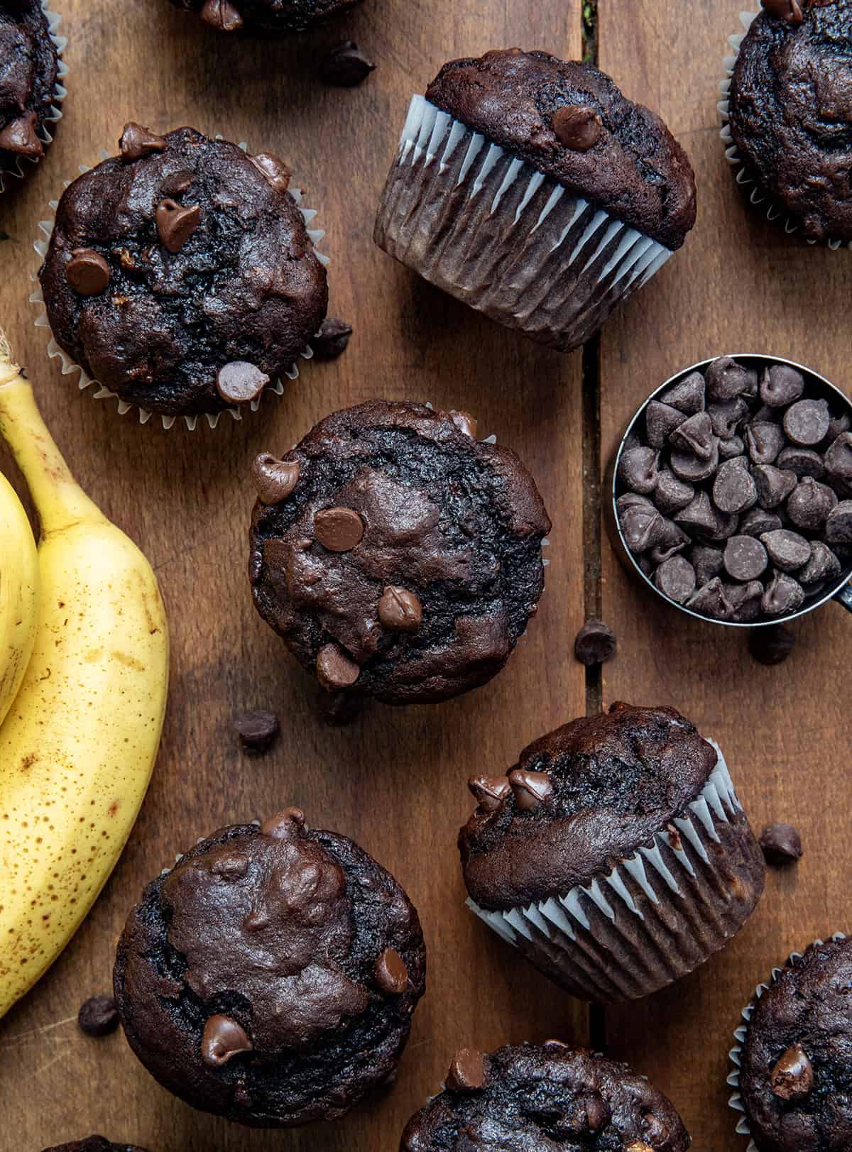 If you are looking for an easy, delicious treat that combines the rich goodness of chocolate with the natural sweetness of ripe bananas, these Double Chocolate Banana Muffins are exactly what you need. (Yes, need!) Double Chocolate Banana Muffins on a wooden table with chocolate chips and bananas from overhead.