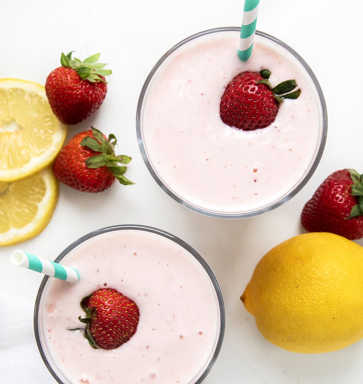 I'm all about beating the heat with this super simple, two-ingredient Frosted Strawberry Lemonade, the ultimate refreshing drink for hot summer days! This tangy 2-ingredient treat comes together in seconds and is perfect for kids and adults alike! Two glasses of Frosted Strawberry Lemonade on a white counter with fresh lemons and strawberries.