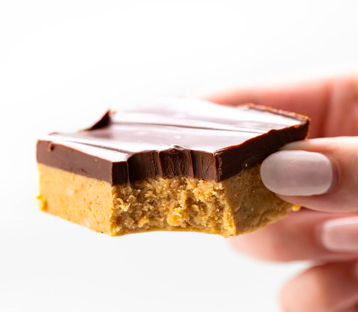 Lunch Lady Peanut Butter Bars are a no-bake dessert with a crunchy graham cracker and peanut butter base topped with a smooth chocolate peanut butter topping. The bars set up in the refrigerator for an easy, delicious treat you can slice and enjoy anytime. Hand holding a bit into Lunch Lady Peanut Butter Bar.
