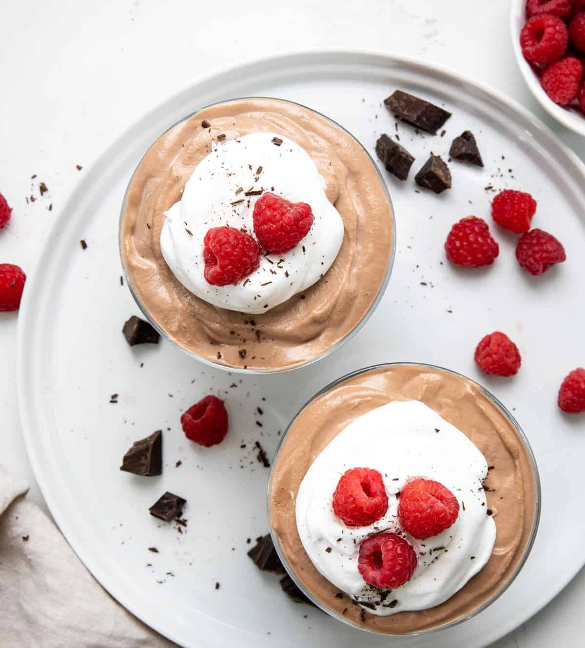 This Two-Ingredient Nutella Mousse is as easy as it gets, and so delicious, you'd never guess it only takes minutes to make! No baking, no eggs, no fuss, just a creamy, dreamy no-bake dessert that feels a little fancy without any extra work. It’s perfect for a quick treat or an easy make-ahead dessert for guests. I like to top mine with a dollop of whipped cream, or even a drizzle of Nutella! Two cups of Nutella Mousse on a white platter with fresh raspberries and shaved chocolate from overhead.