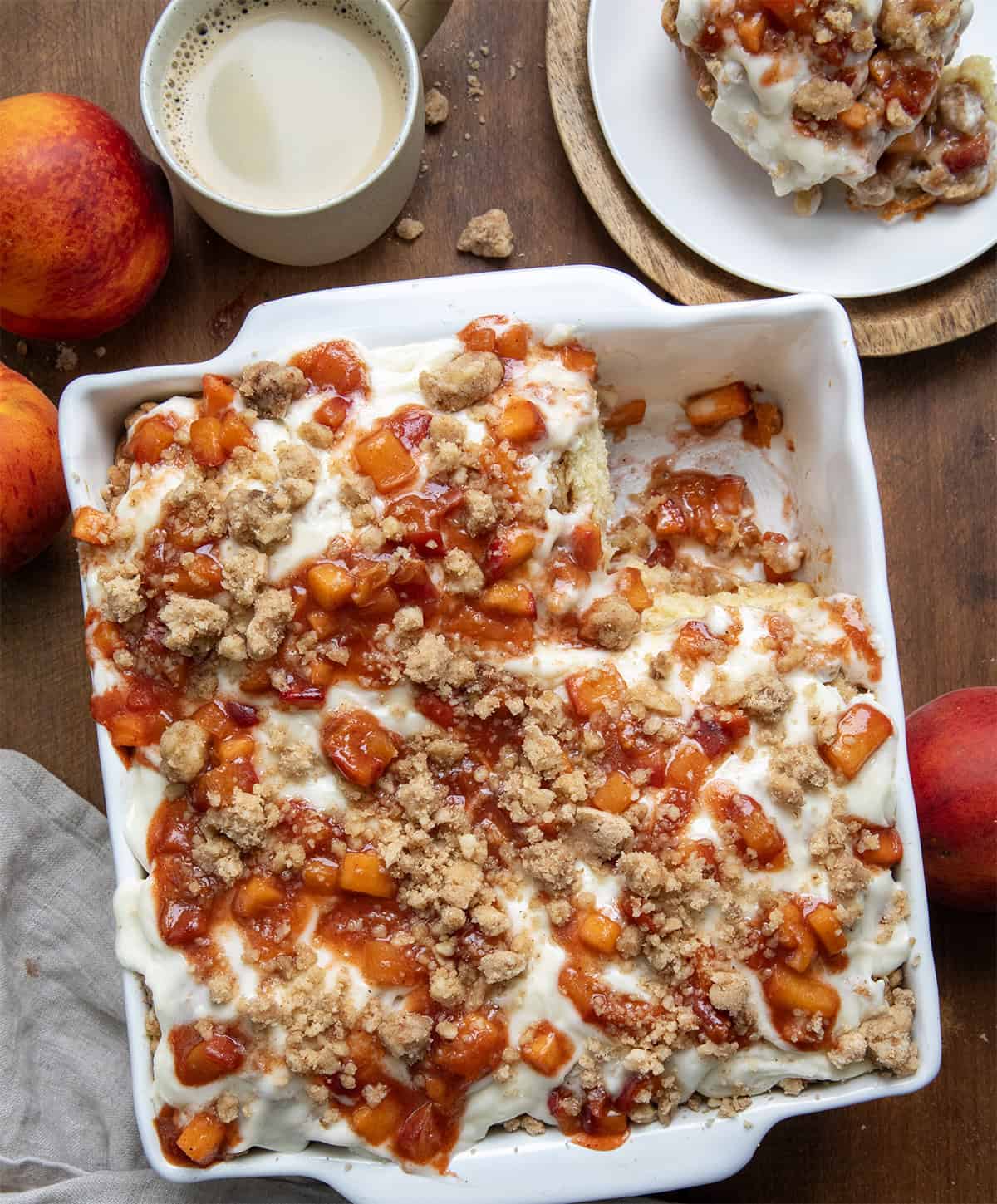 These Peach Cobbler Cinnamon Rolls are the ultimate summer breakfast treat. Soft and fluffy homemade cinnamon rolls are filled with cinnamon-sugar and a juicy fresh peach filling, then topped with a buttery crumble and whipped cream cheese frosting. It’s everything you love about peach cobbler baked into a warm, gooey cinnamon roll! Pan of Peach Cobbler Cinnamon Rolls with one roll removed and on a white plate all on a wooden table from overhead.