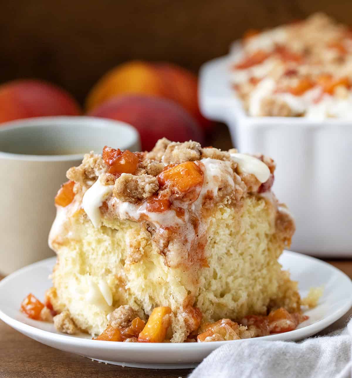 These Peach Cobbler Cinnamon Rolls are the ultimate summer breakfast treat. Soft and fluffy homemade cinnamon rolls are filled with cinnamon-sugar and a juicy fresh peach filling, then topped with a buttery crumble and whipped cream cheese frosting. It’s everything you love about peach cobbler baked into a warm, gooey cinnamon roll! Peach Cobbler Cinnamon Roll on a white plate on a wooden table close up with fresh peaches in the background.