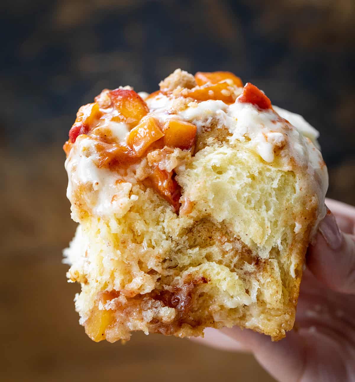 These Peach Cobbler Cinnamon Rolls are the ultimate summer breakfast treat. Soft and fluffy homemade cinnamon rolls are filled with cinnamon-sugar and a juicy fresh peach filling, then topped with a buttery crumble and whipped cream cheese frosting. It’s everything you love about peach cobbler baked into a warm, gooey cinnamon roll! Hand holding a mile-high Peach Cobbler Cinnamon Roll.