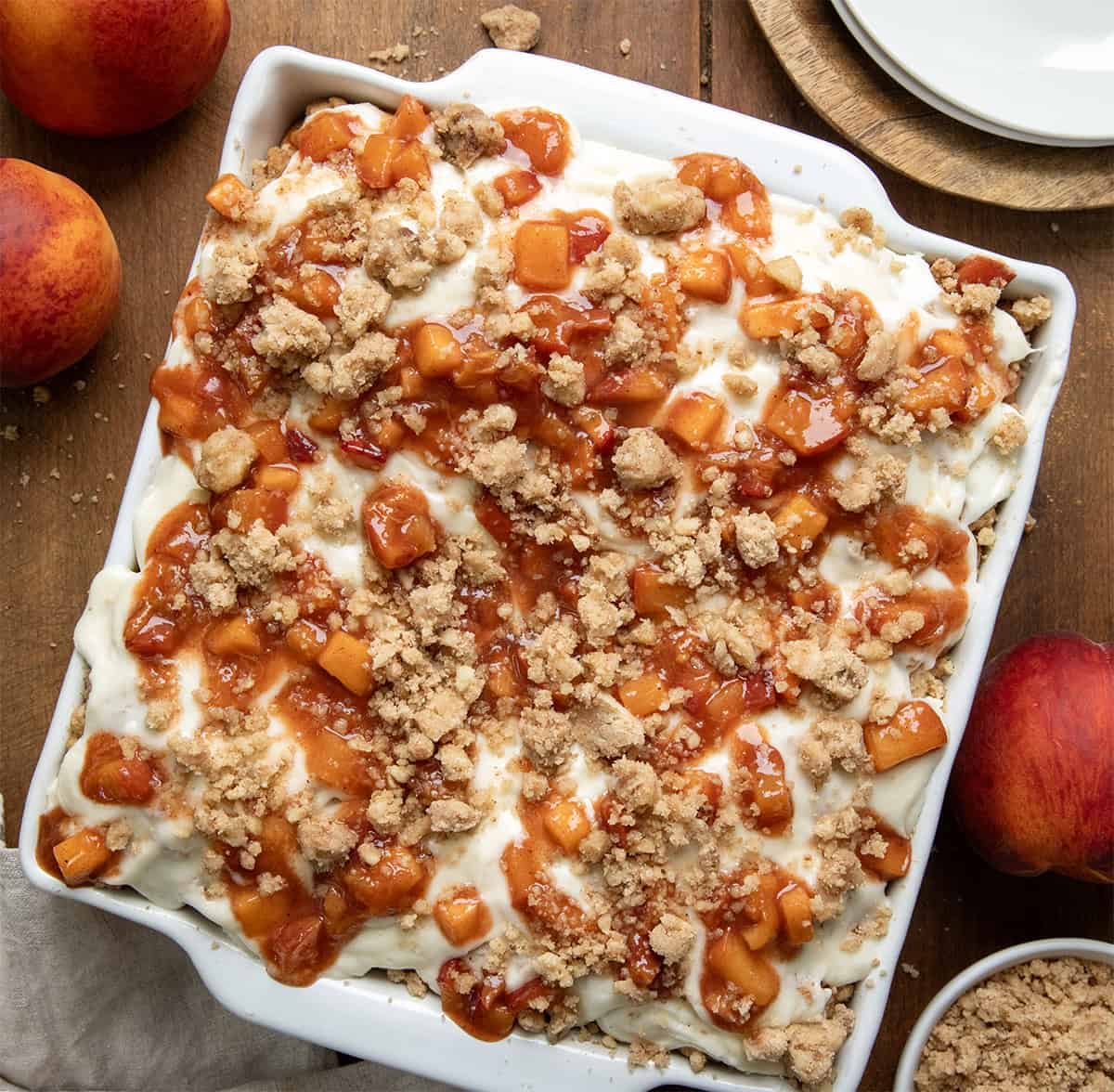 These Peach Cobbler Cinnamon Rolls are the ultimate summer breakfast treat. Soft and fluffy homemade cinnamon rolls are filled with cinnamon-sugar and a juicy fresh peach filling, then topped with a buttery crumble and whipped cream cheese frosting. It’s everything you love about peach cobbler baked into a warm, gooey cinnamon roll! Pan of Peach Cobbler Cinnamon Rolls on a wooden table with fresh peaches from overhead.