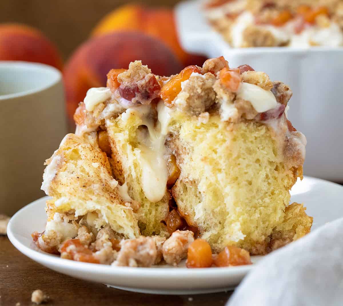 These Peach Cobbler Cinnamon Rolls are the ultimate summer breakfast treat. Soft and fluffy homemade cinnamon rolls are filled with cinnamon-sugar and a juicy fresh peach filling, then topped with a buttery crumble and whipped cream cheese frosting. It’s everything you love about peach cobbler baked into a warm, gooey cinnamon roll! Inside of a Peach Cobbler Cinnamon Roll on a white plate with cream cheese frosting dripping down the roll.