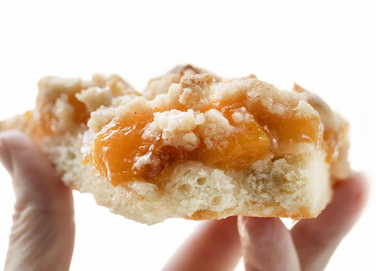 Anytime I see a dessert pizza like Cactus Bread, I want to skip the meal and go straight for the sweet stuff! This Peach Cobbler Pizza starts with my soft, fluffy focaccia bread. On top, you get sweet peaches, a buttery cinnamon crumble, and a drizzle of vanilla glaze. You wonโt want to share! Hand holding a piece of Peach Cobbler Pizza.