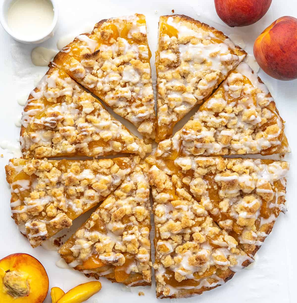 Anytime I see a dessert pizza like Cactus Bread, I want to skip the meal and go straight for the sweet stuff! This Peach Cobbler Pizza starts with my soft, fluffy focaccia bread. On top, you get sweet peaches, a buttery cinnamon crumble, and a drizzle of vanilla glaze. You wonโt want to share! Peach Cobbler Pizza cut into slices on a white table.