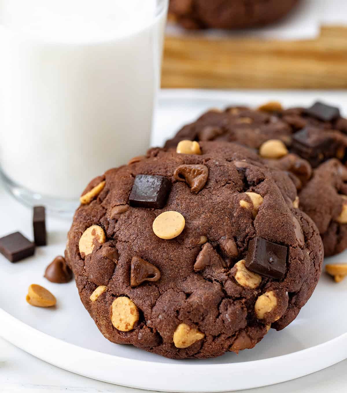 These Peanut Butter Chocolate Chunk Cookies are extra large, extra thick, bakery-style chocolate cookies loaded with semi-sweet chocolate, milk chocolate, and peanut butter chips! Peanut Butter Chocolate Chunk Cookies on a white plate with a glass of milk close up.